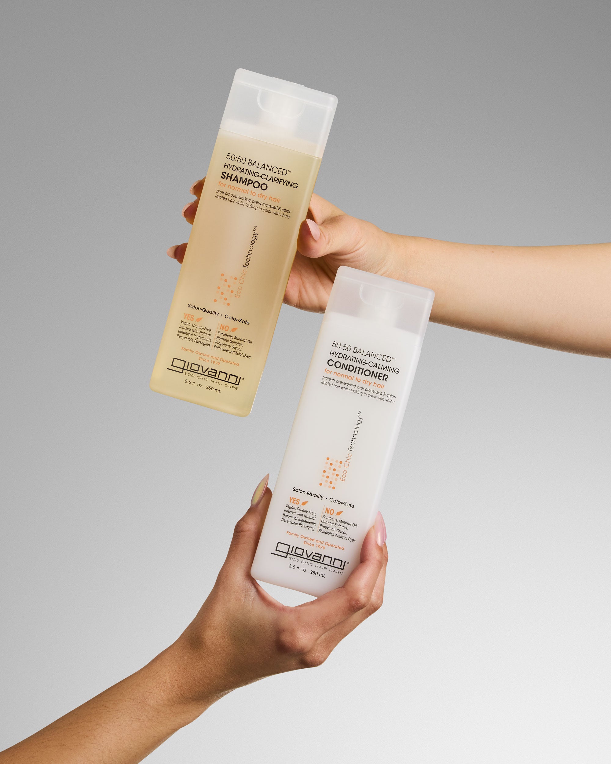 Two hands hold Giovanni Cosmetics hair care bottles against a gray background—one is 50:50 Balanced Hydrating-Clarifying Shampoo, the other is 50:50 BALANCED™ HYDRATING-CALMING CONDITIONER.
