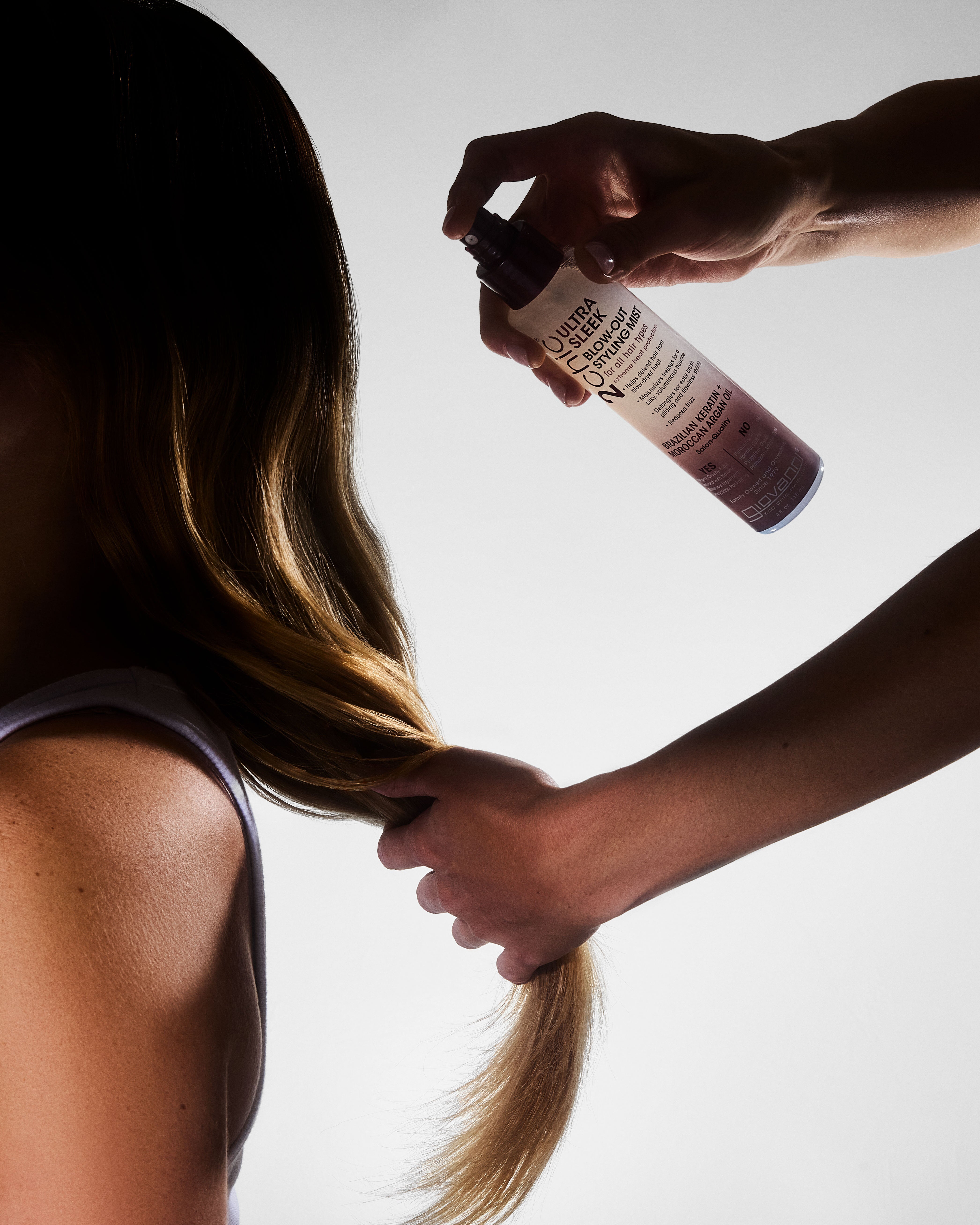 Person applying 2Chic Ultra Sleek Blow-Out Styling Mist to another person's hair with a white background