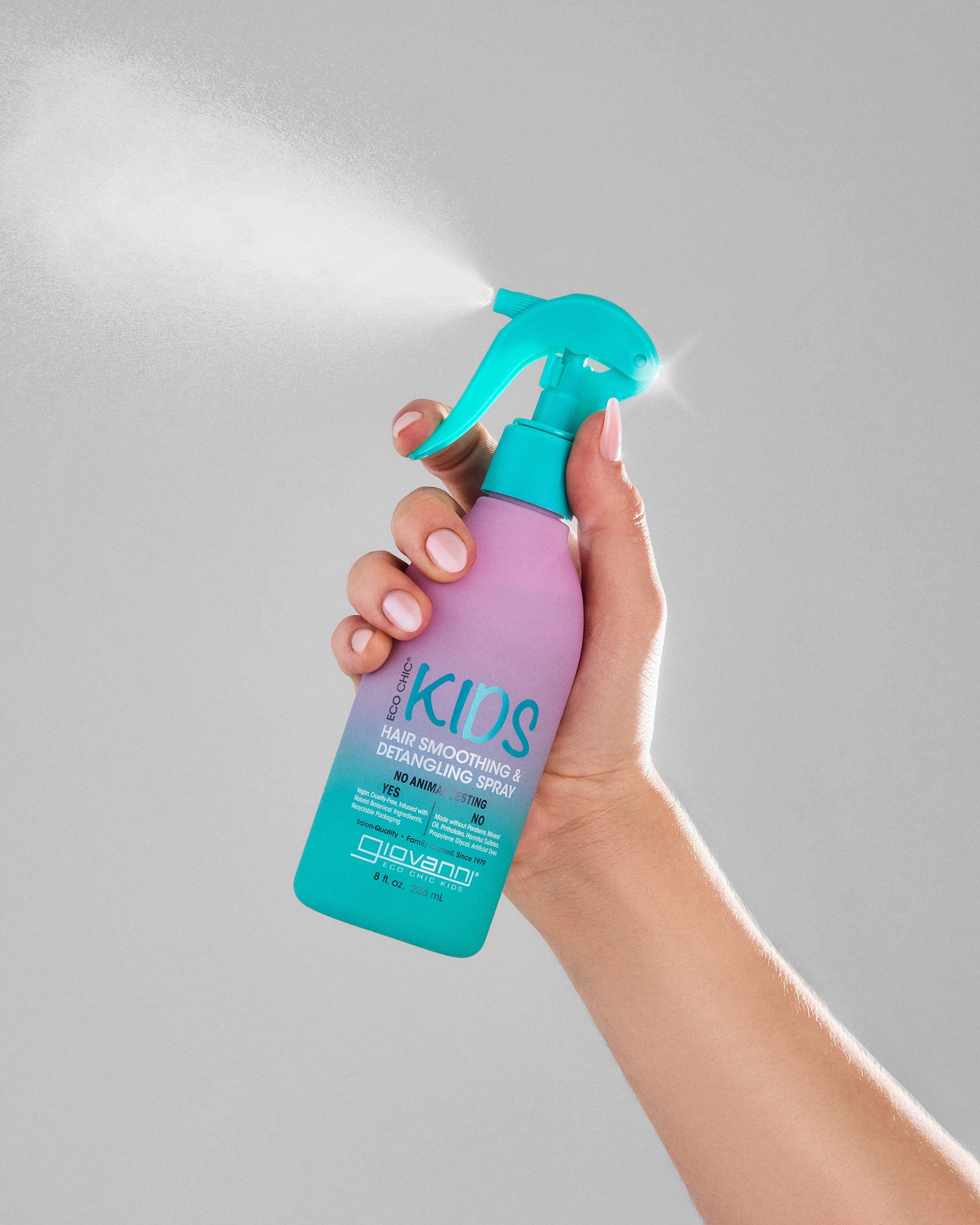 Hand holding Giovanni Eco Chic Kids Hair Smoothing & Detangling Spray bottle against a gray background