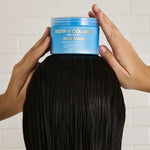 Person holding a jar of Giovanni Biotin & Collagen hair mask over long black hair against a white tiled wall.