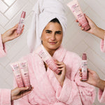 Woman in a pink robe holding a bottle of 
Giovanni 2Chic Frizz Be Gone Shampoo surrounded by hands holding other Frizz Be Gone products