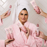 Woman in a pink robe holding a bottle of 
Giovanni 2Chic Frizz Be Gone Shampoo surrounded by hands holding other Frizz Be Gone products
