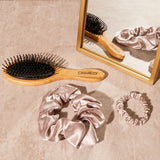A set of three Giovanni Cosmetics SATIN HAIR SCRUNCHIES in Blush, Gray, and Black is displayed alongside a wooden hairbrush on a beige surface near a gold-framed mirror reflecting the elegant hair accessories.
