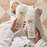 Person wrapped in a towel holding Giovanni Tea Tree Triple Treat shampoo and conditioner bottles.