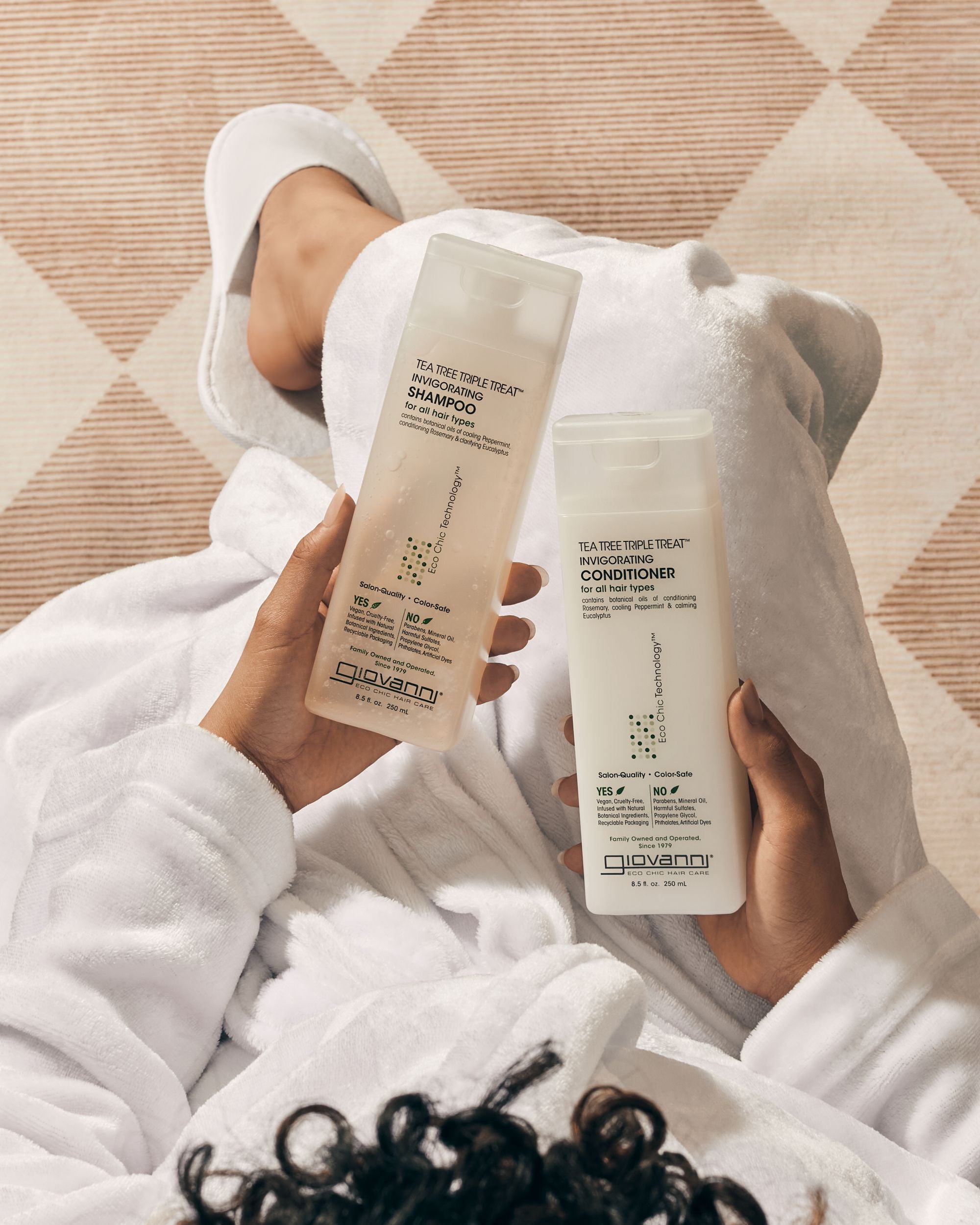 Person wrapped in a towel holding Giovanni Tea Tree Triple Treat shampoo and conditioner bottles.