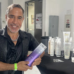 A person smiles and holds a bottle of Giovanni Cosmetics COLOR CHIC™ PURPLE SHAMPOO at a display table with various hair and beauty products arranged neatly in a well-lit indoor setting, ready to banish brassy tones for brighter blonde hair.