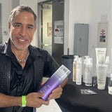A person smiles and holds a bottle of Giovanni Cosmetics COLOR CHIC™ PURPLE SHAMPOO at a display table with various hair and beauty products arranged neatly in a well-lit indoor setting, ready to banish brassy tones for brighter blonde hair.