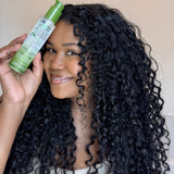 Woman with curly hair holding a bottle of 2Chic Ultra Moist serum against a neutral background