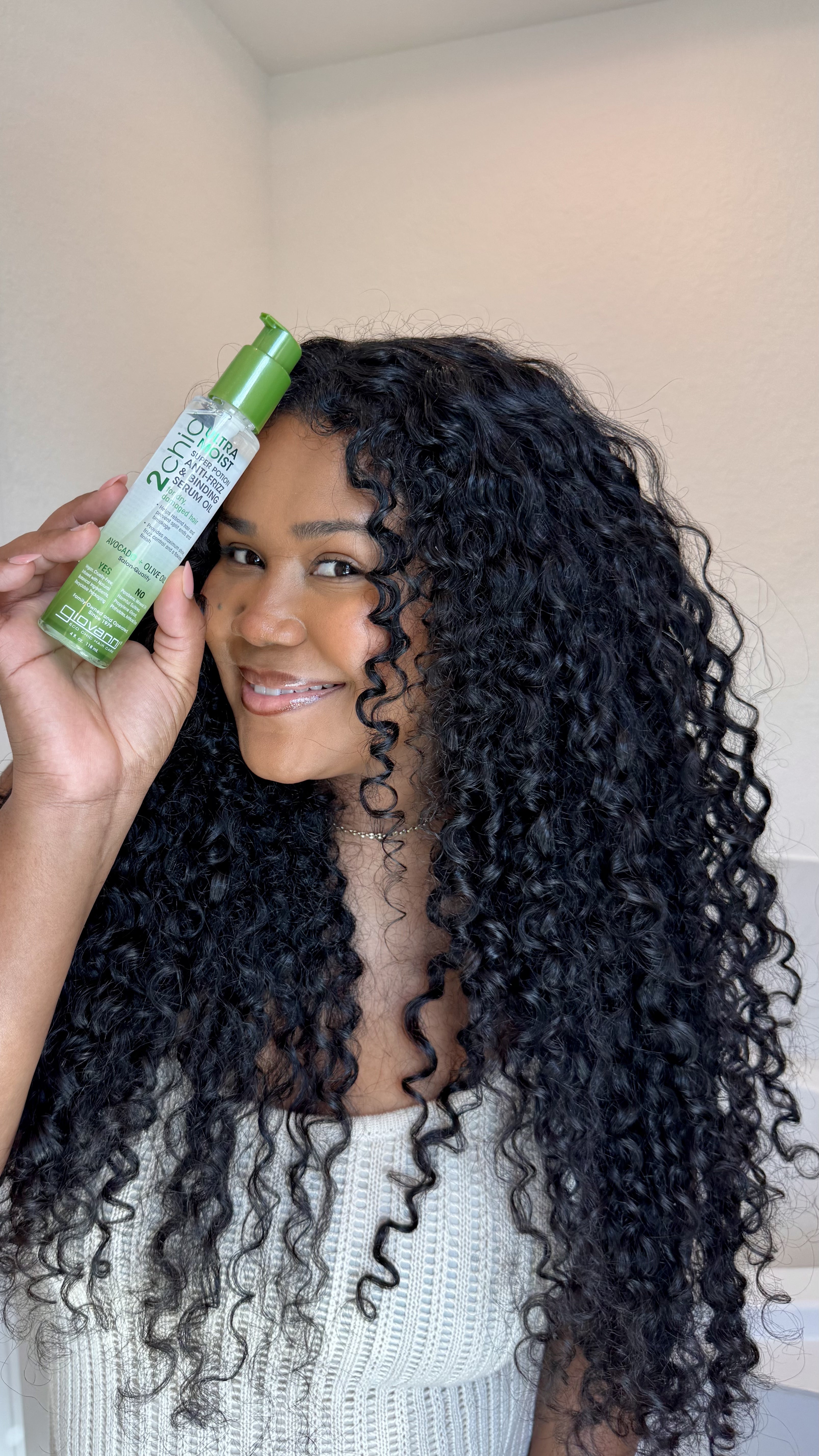 Woman with curly hair holding a bottle of 2Chic Ultra Moist serum against a neutral background