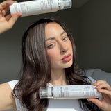 A woman with long, wavy brown hair and a white shirt holds two Giovanni Cosmetics SMOOTH AS SILK™ HAIR DETANGLING SPRAY bottles—one above her head, one near her chin. Her silky hair shines in soft natural light against a neutral background.