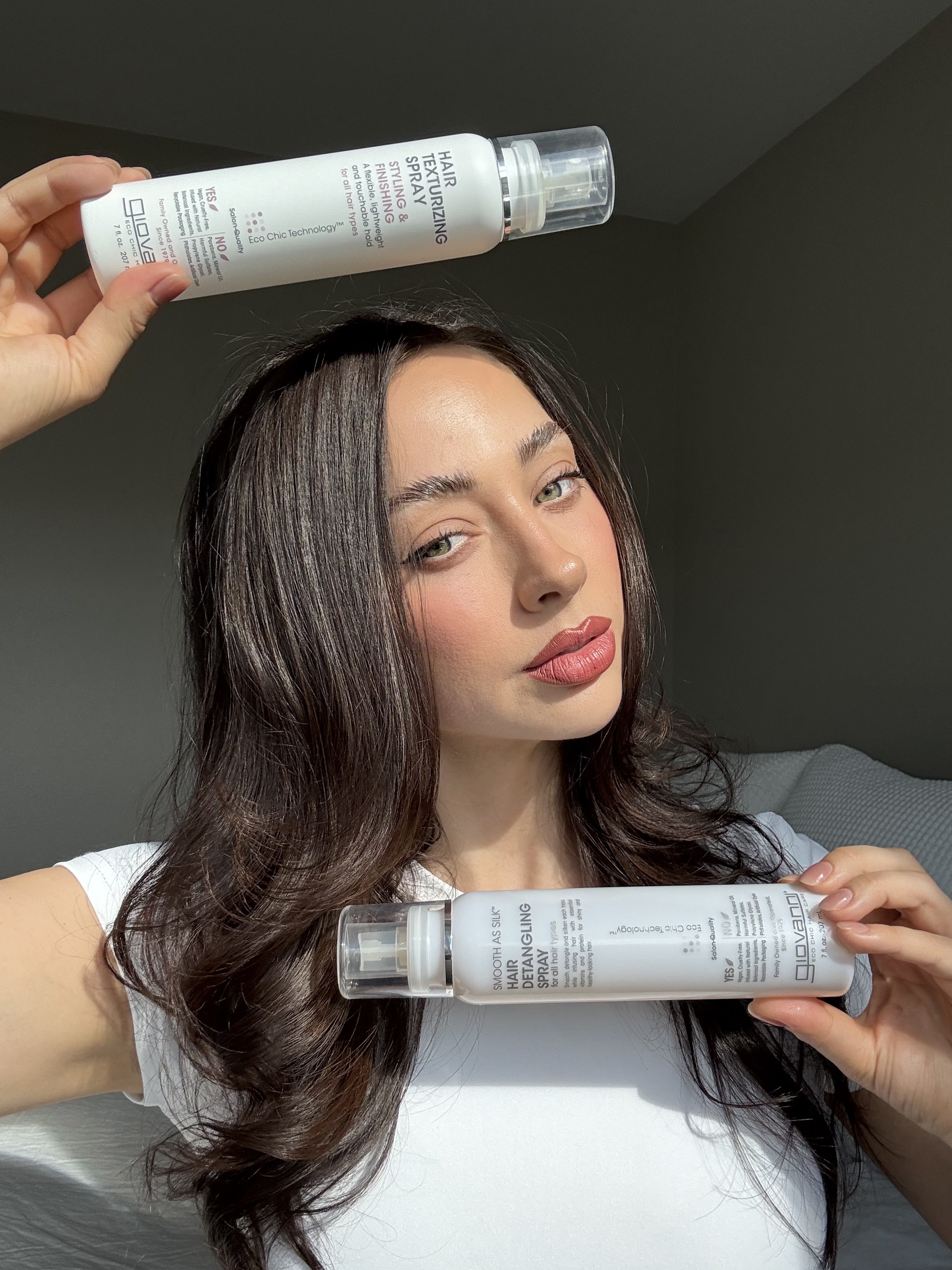 A woman with long, wavy brown hair and a white shirt holds two Giovanni Cosmetics SMOOTH AS SILK™ HAIR DETANGLING SPRAY bottles—one above her head, one near her chin. Her silky hair shines in soft natural light against a neutral background.