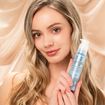 A woman with wavy blonde hair and natural makeup smiles softly, holding a blue bottle of Giovanni Cosmetics BIOTIN & COLLAGEN STRENGTHENING SMOOTHING & STYLING HAIR SERUM against a silky beige backdrop.