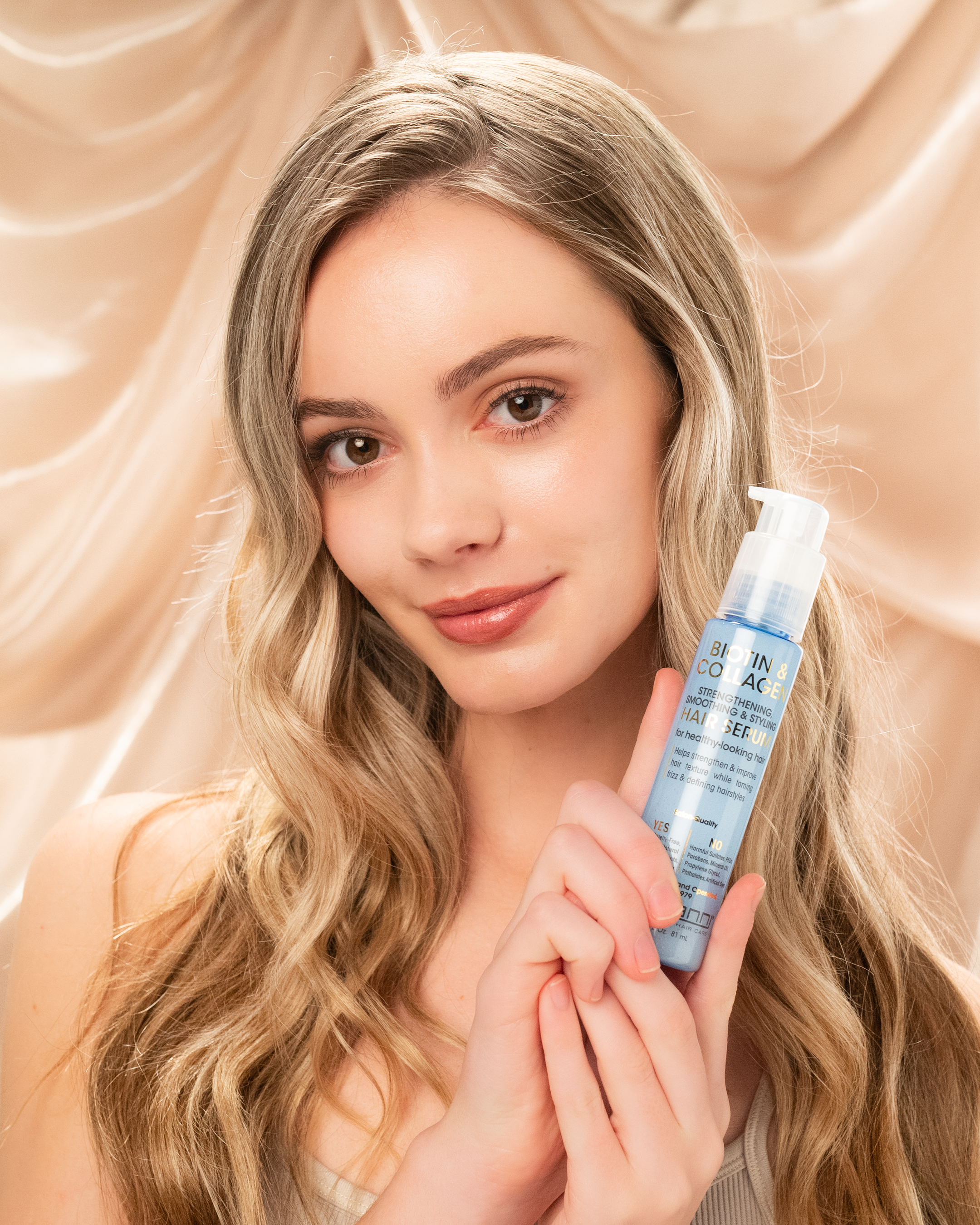 A woman with wavy blonde hair and natural makeup smiles softly, holding a blue bottle of Giovanni Cosmetics BIOTIN & COLLAGEN STRENGTHENING SMOOTHING & STYLING HAIR SERUM against a silky beige backdrop.