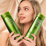 A smiling woman with long, wavy blonde hair holds two green bottles of Giovanni Cosmetics HEMP HYDRATING SHAMPOO in front of a beige, draped fabric background.