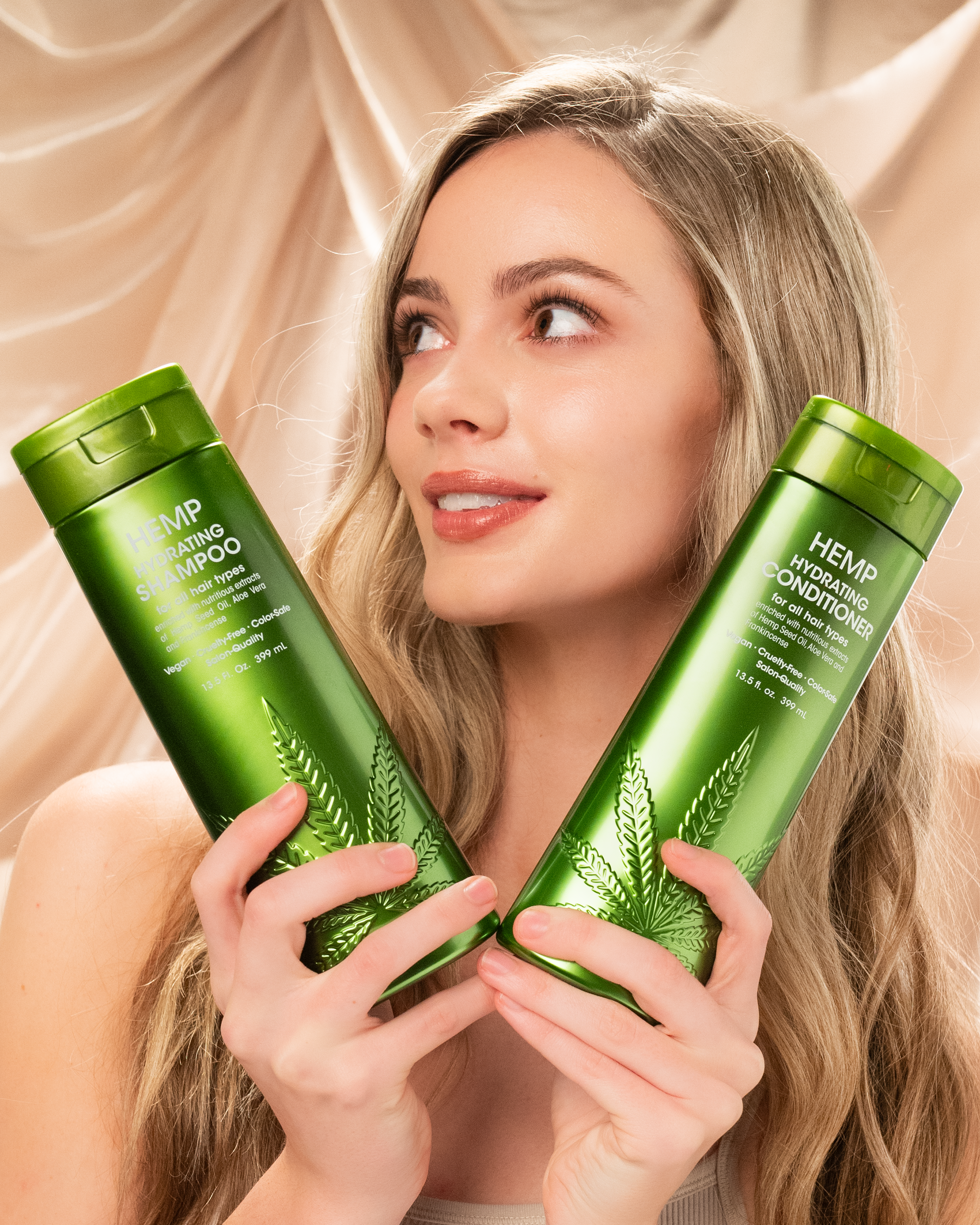 A smiling woman with long, wavy blonde hair holds two green bottles of Giovanni Cosmetics HEMP HYDRATING SHAMPOO in front of a beige, draped fabric background.