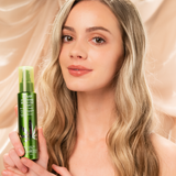 A woman with long, wavy blonde hair smiles gently while holding a bottle of Giovanni Cosmetics HEMP HYDRATING BLOW-OUT SPRAY against a beige, silky background.