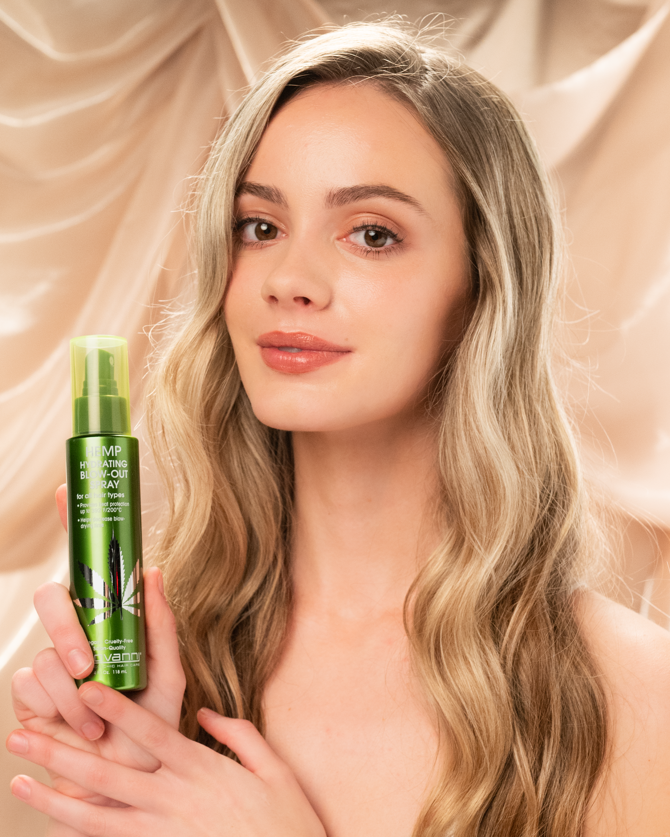 A woman with long, wavy blonde hair smiles gently while holding a bottle of Giovanni Cosmetics HEMP HYDRATING BLOW-OUT SPRAY against a beige, silky background.