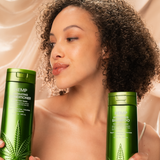 A woman with curly hair smiles softly, holding green bottles of Giovanni Cosmetics HEMP HYDRATING CONDITIONER against a silky beige background. The salon-quality formula features stylish hemp leaf designs and is available in 3 sizes.
