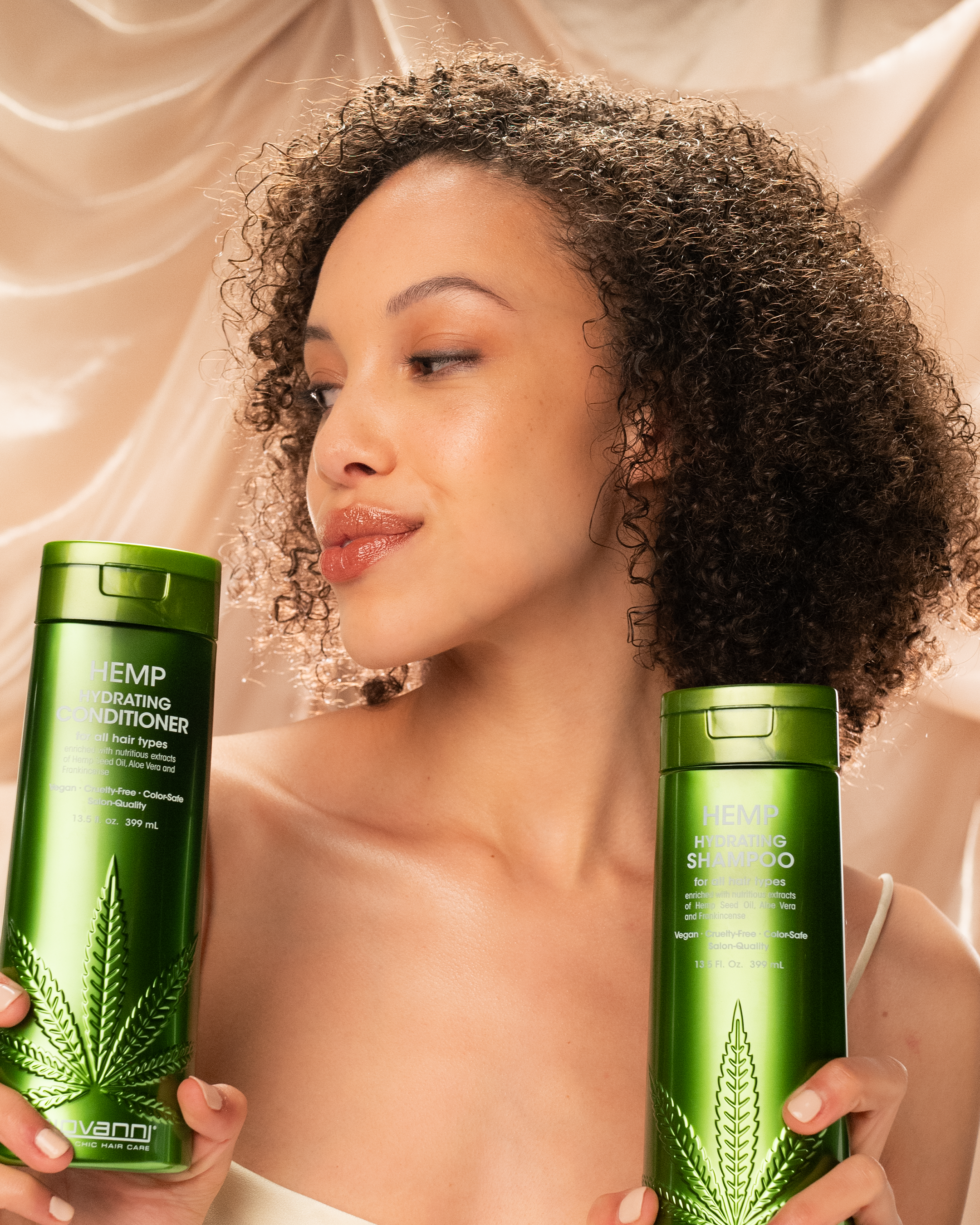 A woman with curly hair smiles softly, holding green bottles of Giovanni Cosmetics HEMP HYDRATING CONDITIONER against a silky beige background. The salon-quality formula features stylish hemp leaf designs and is available in 3 sizes.