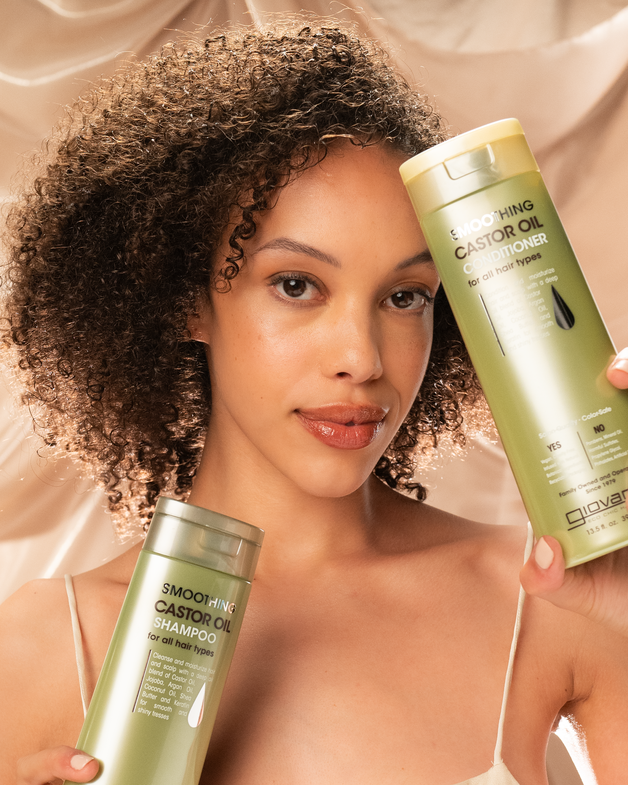 A woman with curly hair smiles softly, holding Giovanni Cosmetics Smoothing Castor Oil Conditioner in front of a beige, silky background—highlighting her frizz-taming and hair protection routine.