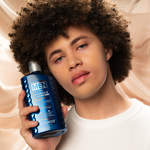 A person with curly hair holds Giovanni Cosmetics MEN 2-in-1 Daily Shampoo & Conditioner with Ginseng & Eucalyptus, highlighting natural men’s hair care against a soft beige fabric background.
