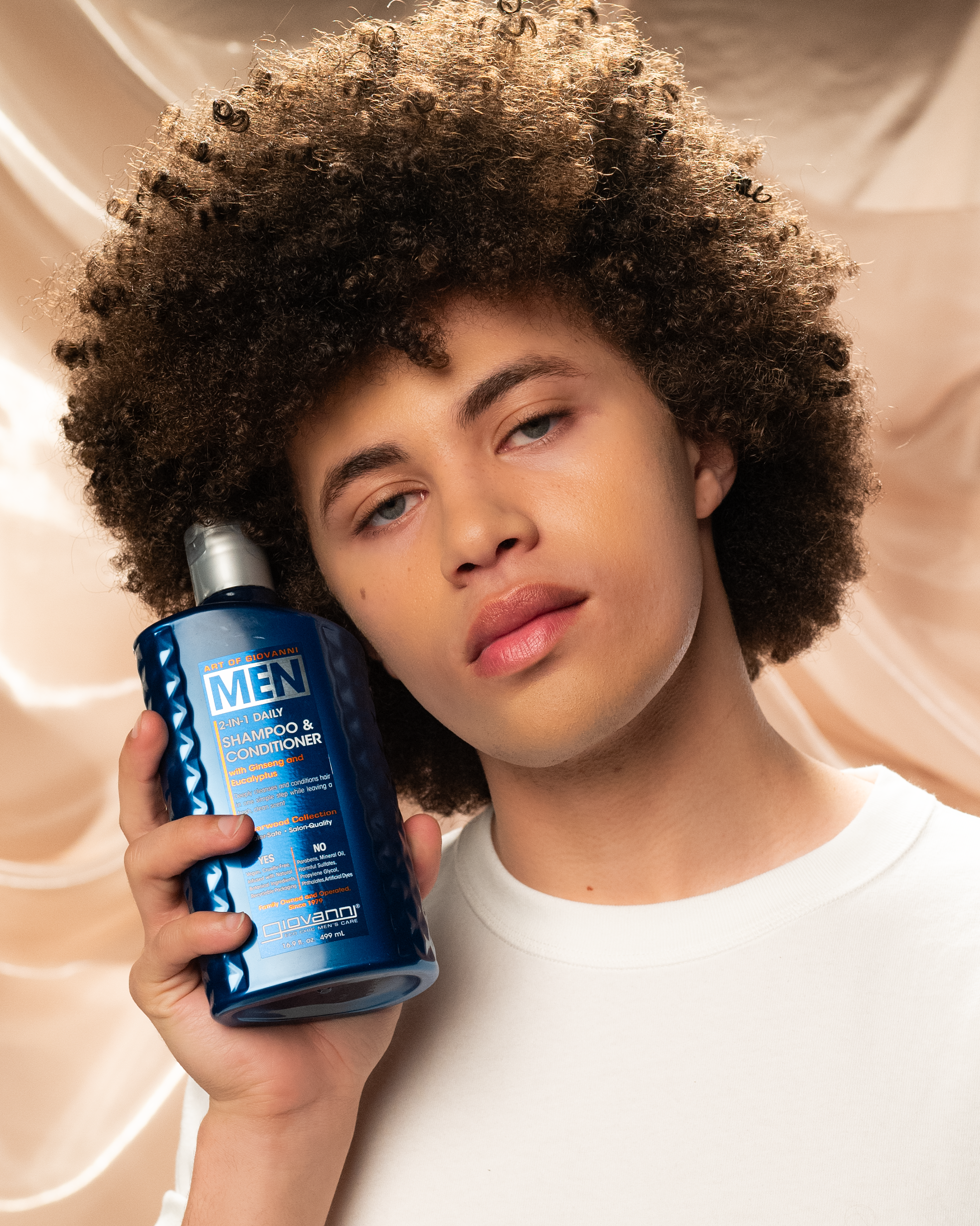A person with curly hair holds Giovanni Cosmetics MEN 2-in-1 Daily Shampoo & Conditioner with Ginseng & Eucalyptus, highlighting natural men’s hair care against a soft beige fabric background.