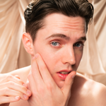 A young man with fair skin and dark hair touches his cheek, looking concerned—perhaps in need of Giovanni Cosmetics' MEN Moisturizing Shave Cream. Soft beige fabric drapes in the background.