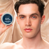 A young man with light skin and brown hair holds up a blue container of MEN Styling Hair Clay by Giovanni Cosmetics, known for its strong hold, looking directly at the camera with a neutral expression.