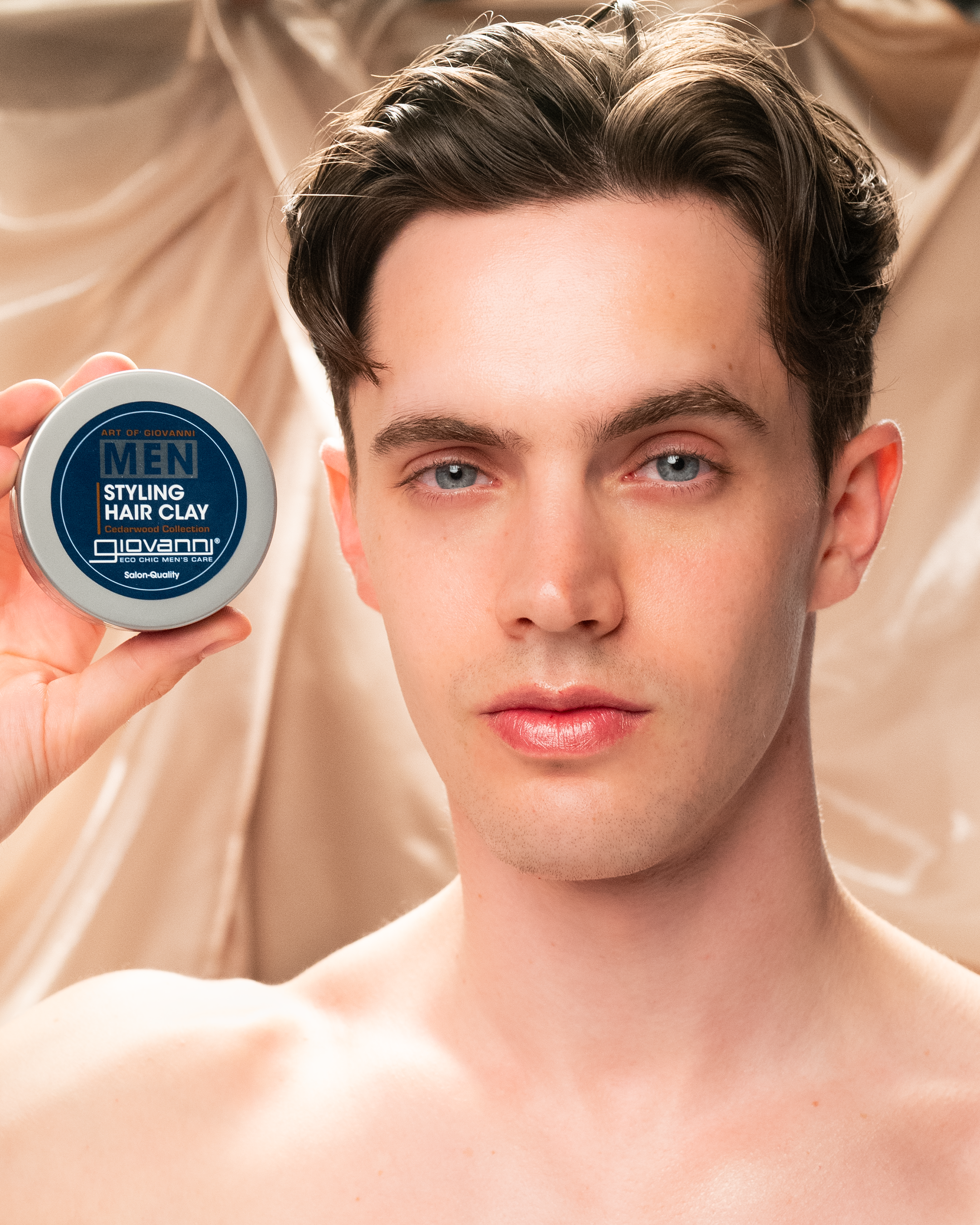 A young man with light skin and brown hair holds up a blue container of MEN Styling Hair Clay by Giovanni Cosmetics, known for its strong hold, looking directly at the camera with a neutral expression.