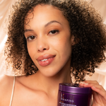 A woman with curly hair smiles at the camera, holding a purple container of Giovanni Cosmetics CURL HABIT™ CURL SHAPING HAIR GEL. The soft, beige silky background highlights this curl-defining gel infused with moisturizing oils.