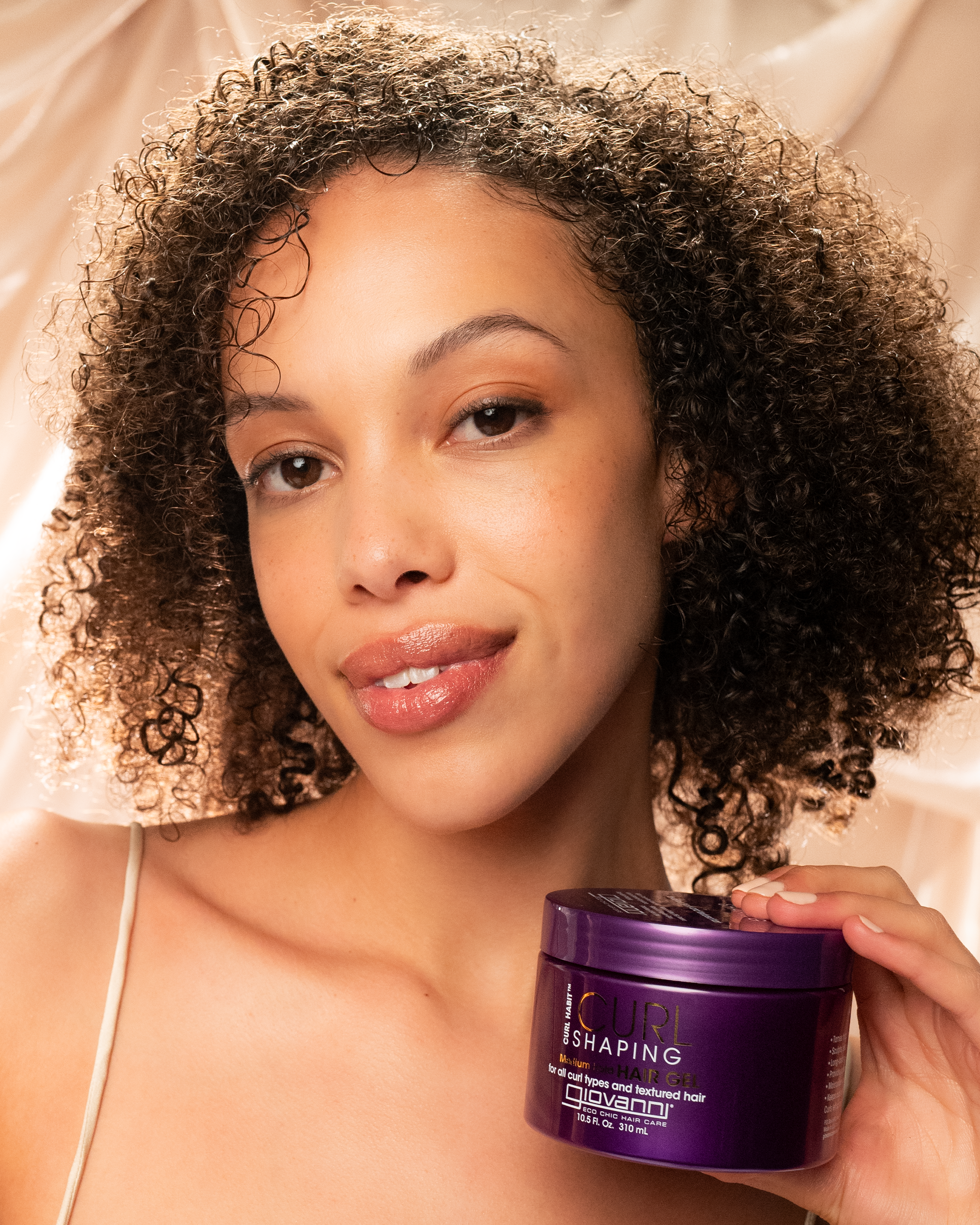 A woman with curly hair smiles at the camera, holding a purple container of Giovanni Cosmetics CURL HABIT™ CURL SHAPING HAIR GEL. The soft, beige silky background highlights this curl-defining gel infused with moisturizing oils.