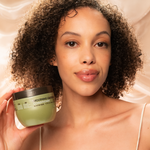 A woman with curly hair smiles softly at the camera, holding up Giovanni Cosmetics SMOOTHING CASTOR OIL LEAVE-IN CONDITIONER against a creamy backdrop—the perfect solution for smooth, defined, frizz-free curls.