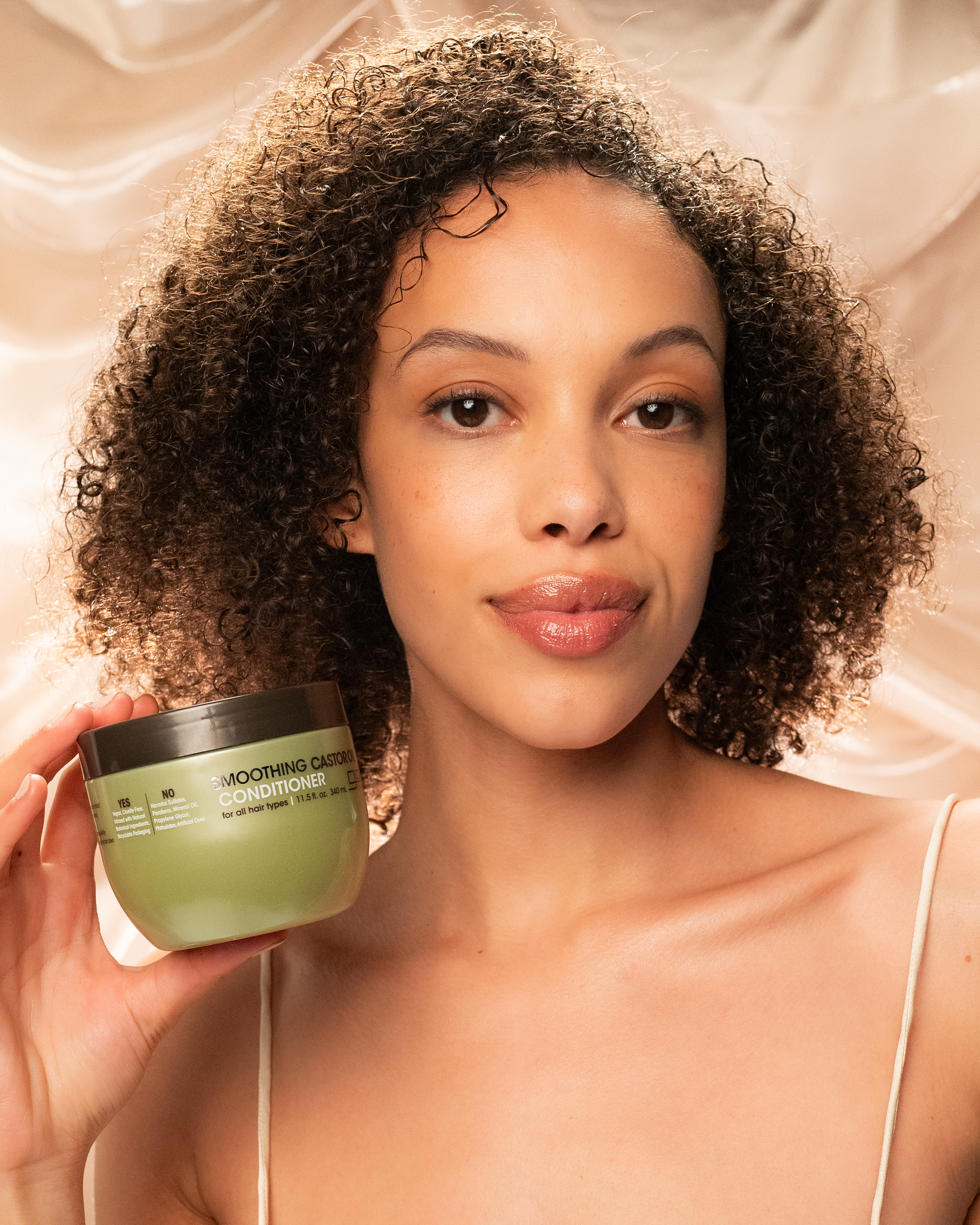 A woman with curly hair smiles softly at the camera, holding up Giovanni Cosmetics SMOOTHING CASTOR OIL LEAVE-IN CONDITIONER against a creamy backdrop—the perfect solution for smooth, defined, frizz-free curls.