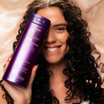 A smiling woman with long, curly hair holds a large bottle of Giovanni Cosmetics CURL HABIT™ CURL DEFINING CONDITIONER—available in 2 sizes—next to her face, ready to define and moisturize curls against a soft beige background.