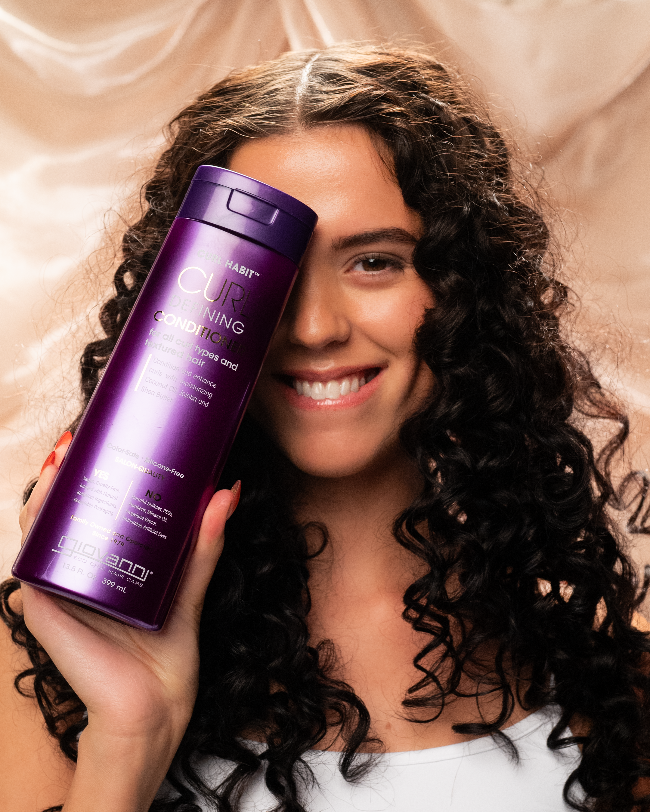 A smiling woman with long, curly hair holds a large bottle of Giovanni Cosmetics CURL HABIT™ CURL DEFINING CONDITIONER—available in 2 sizes—next to her face, ready to define and moisturize curls against a soft beige background.