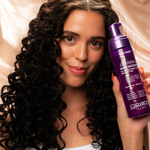 A woman with long, dark curly hair in a white top smiles while holding a purple bottle of Giovanni Cosmetics CURL HABIT™ CURL DEFINING HAIR MOUSSE against a beige satin background.