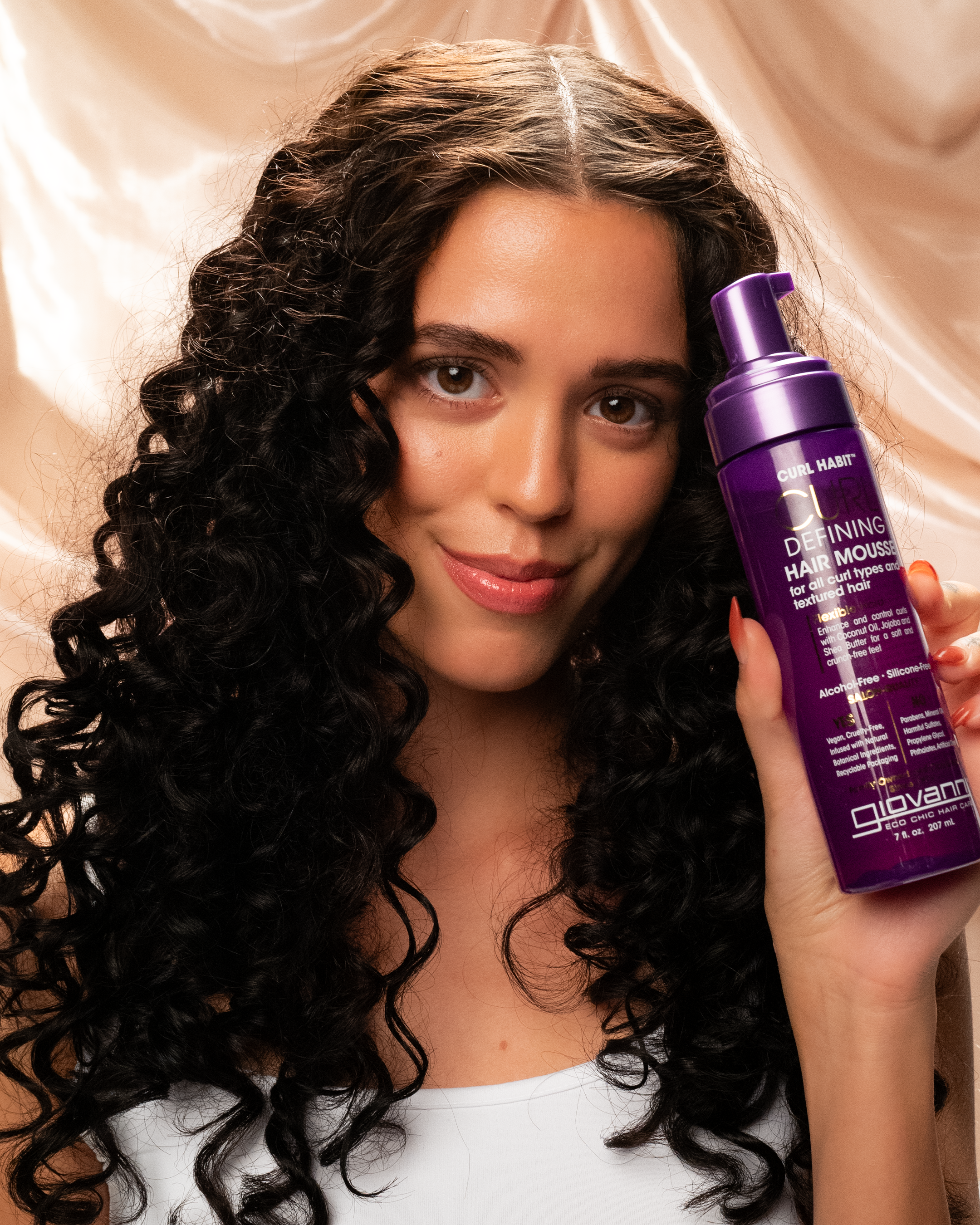 A woman with long, dark curly hair in a white top smiles while holding a purple bottle of Giovanni Cosmetics CURL HABIT™ CURL DEFINING HAIR MOUSSE against a beige satin background.