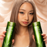 A woman with long, straight, light brown hair holds up green bottles of Giovanni Cosmetics HEMP HYDRATING CONDITIONER in front of a beige satin background. The vegan hair care bottles feature a hemp leaf design and come in 3 sizes.