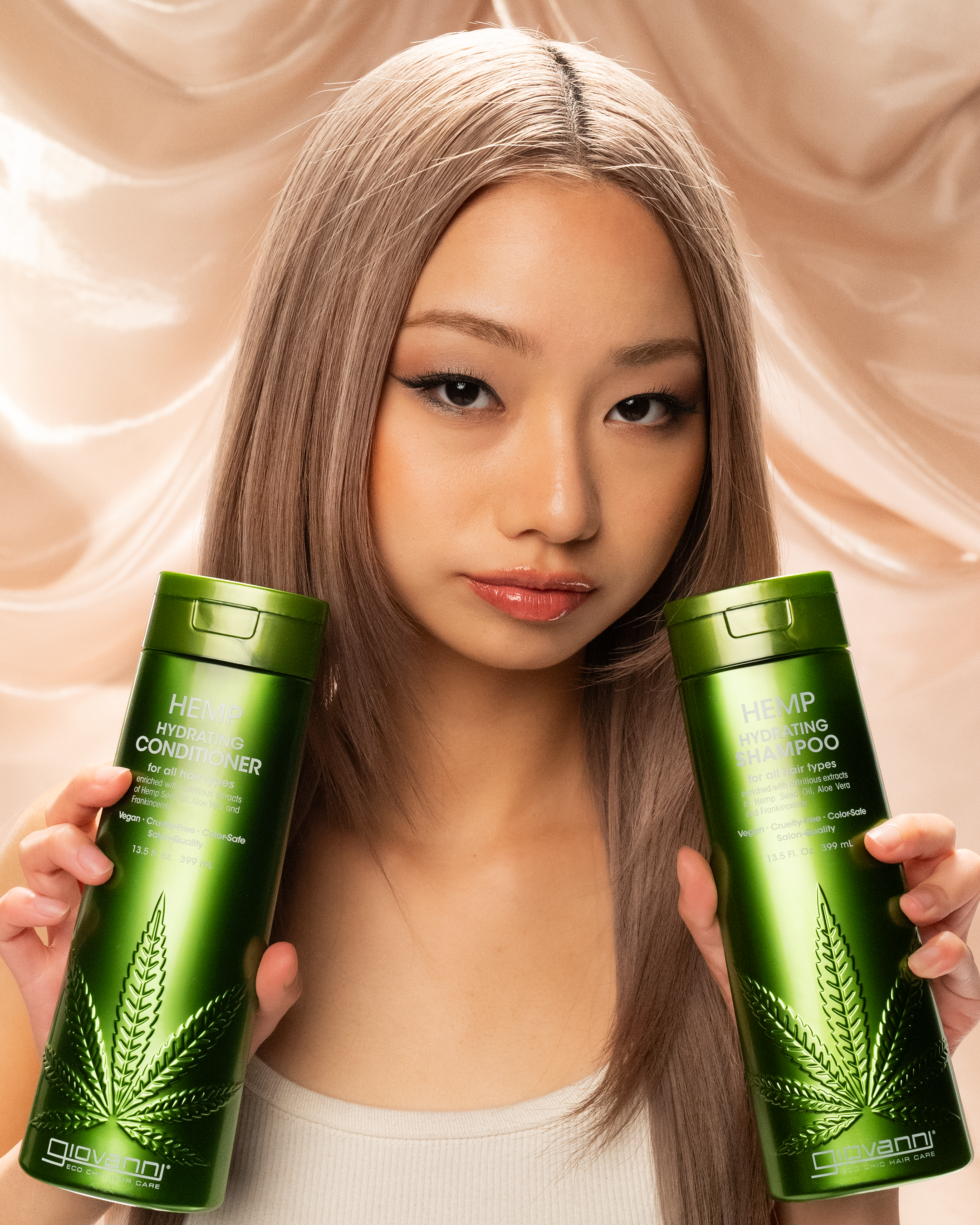 A woman with long, straight, light brown hair holds up green bottles of Giovanni Cosmetics HEMP HYDRATING CONDITIONER in front of a beige satin background. The vegan hair care bottles feature a hemp leaf design and come in 3 sizes.