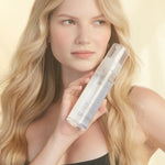 A woman with long blonde hair in a black top holds a clear bottle of Giovanni Cosmetics MOUSSE AIR-TURBO CHARGED™ HAIR STYLING FOAM near her face, posing against a soft, light background.