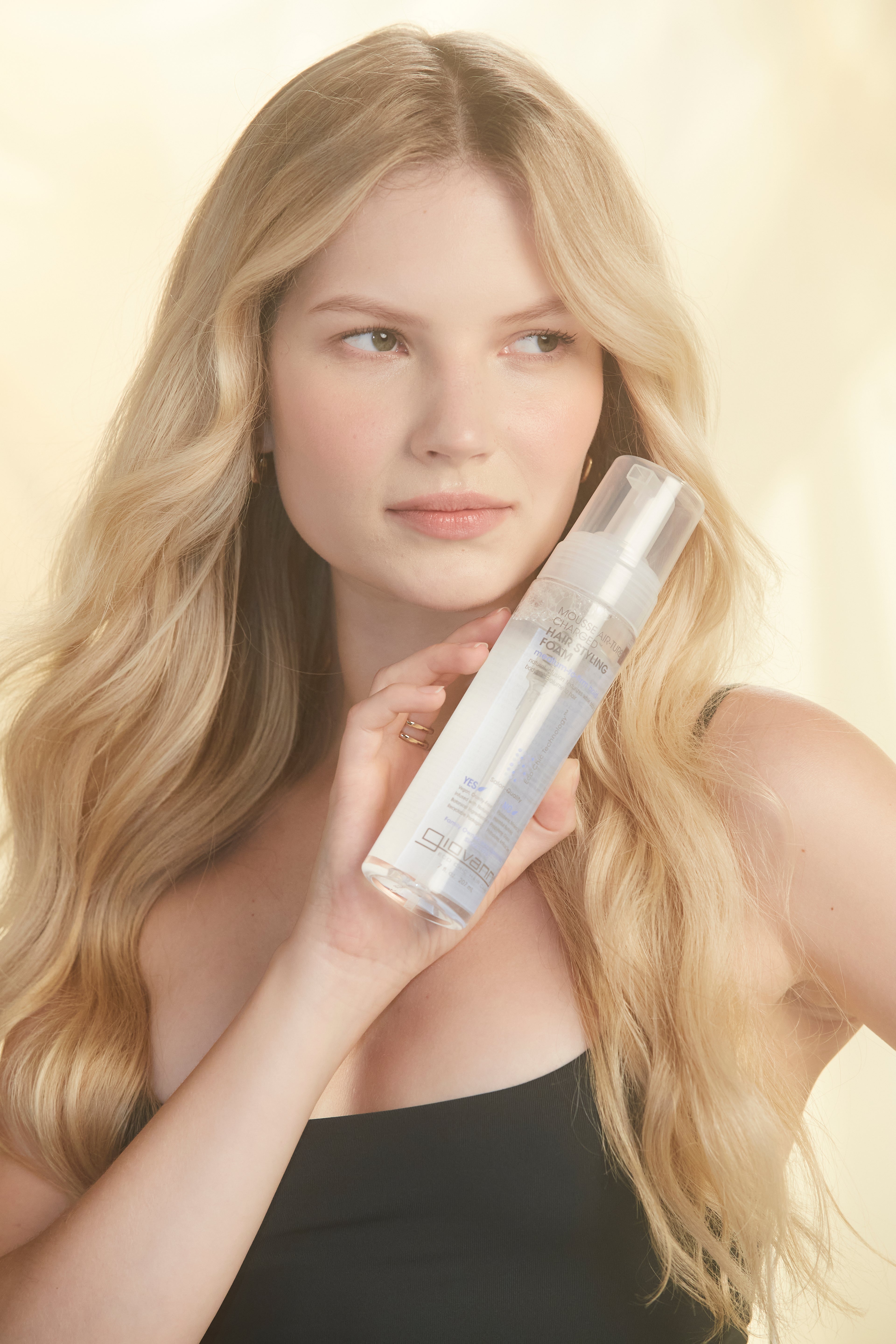 A woman with long blonde hair in a black top holds a clear bottle of Giovanni Cosmetics MOUSSE AIR-TURBO CHARGED™ HAIR STYLING FOAM near her face, posing against a soft, light background.