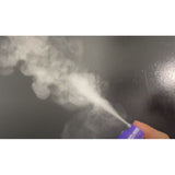 A hand presses the nozzle of a Giovanni Cosmetics POWDER POWER® DRY SHAMPOO bottle, releasing a visible mist against a plain dark background.