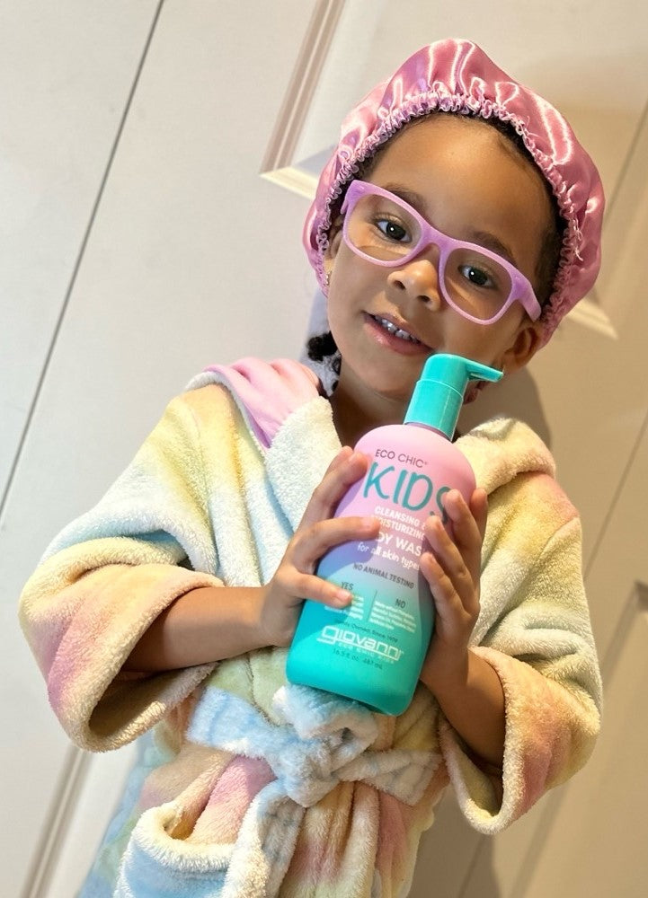 A young child in a pastel bathrobe, pink glasses, and a satin shower cap smiles while holding a large bottle of Giovanni Cosmetics ECO CHIC® KIDS CLEANSING & MOISTURIZING BODY WASH for gentle cleansing and extra moisture.