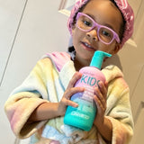 A young child in a pastel bathrobe, pink glasses, and a satin shower cap smiles while holding a large bottle of Giovanni Cosmetics ECO CHIC® KIDS CLEANSING & MOISTURIZING BODY WASH for gentle cleansing and extra moisture.