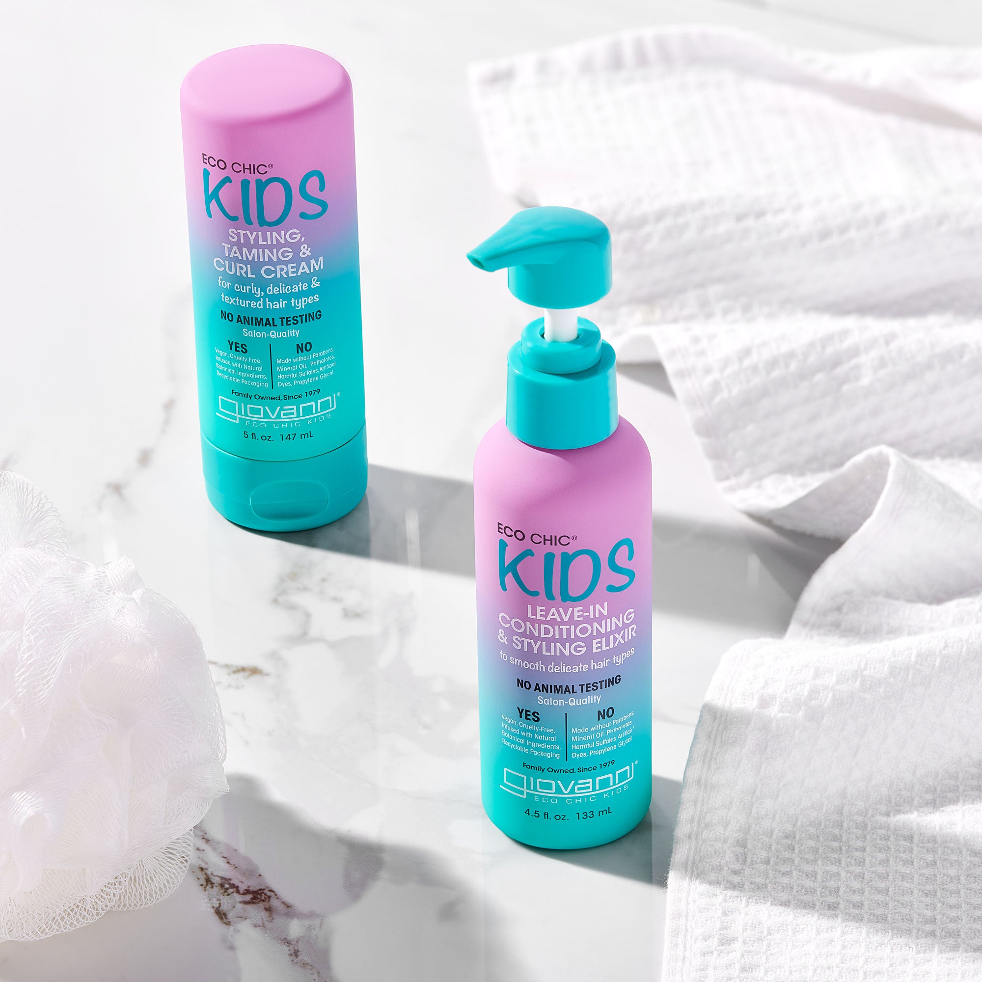Two bottles of Giovanni Cosmetics' ECO CHIC® KIDS STYLING TAMING & CURL CREAM rest on white marble beside towels and a bath sponge, making it easy to care for your child's hair and define every curl.