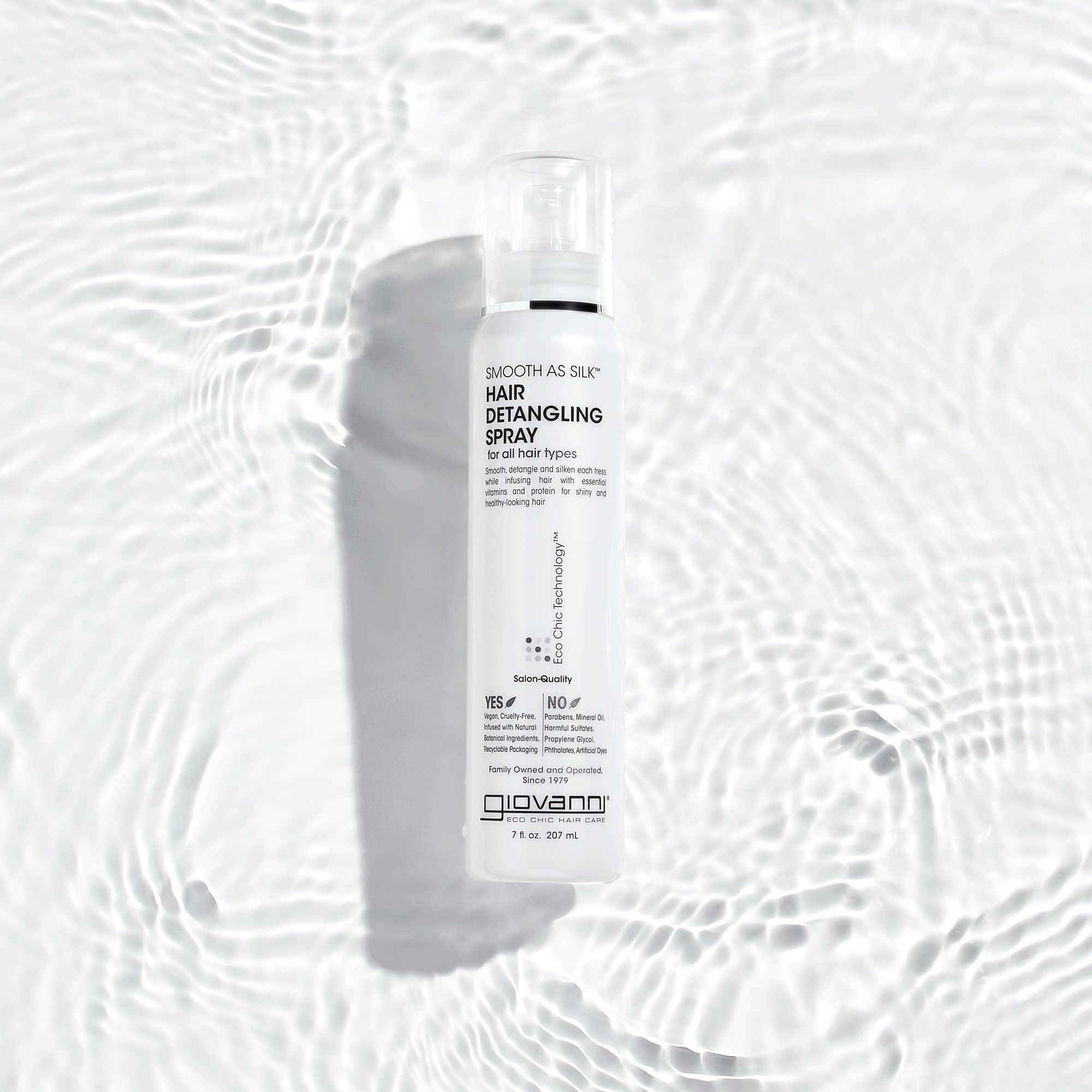 A white bottle of Giovanni Cosmetics SMOOTH AS SILK™ HAIR DETANGLING SPRAY rests on a rippled white surface with soft light reflections, creating a clean, fresh look—ideal for silky, detangled hair.