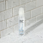 A clear bottle of Giovanni Cosmetics FRIZZ BE GONE™ Super-Smoothing, Anti-Frizz Hair Serum stands upright on a wet, textured surface with water droplets, in front of a white glossy tiled wall with splashes.