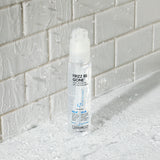 A clear bottle of Giovanni Cosmetics FRIZZ BE GONE™ Super-Smoothing, Anti-Frizz Hair Serum stands upright on a wet, textured surface with water droplets, in front of a white glossy tiled wall with splashes.