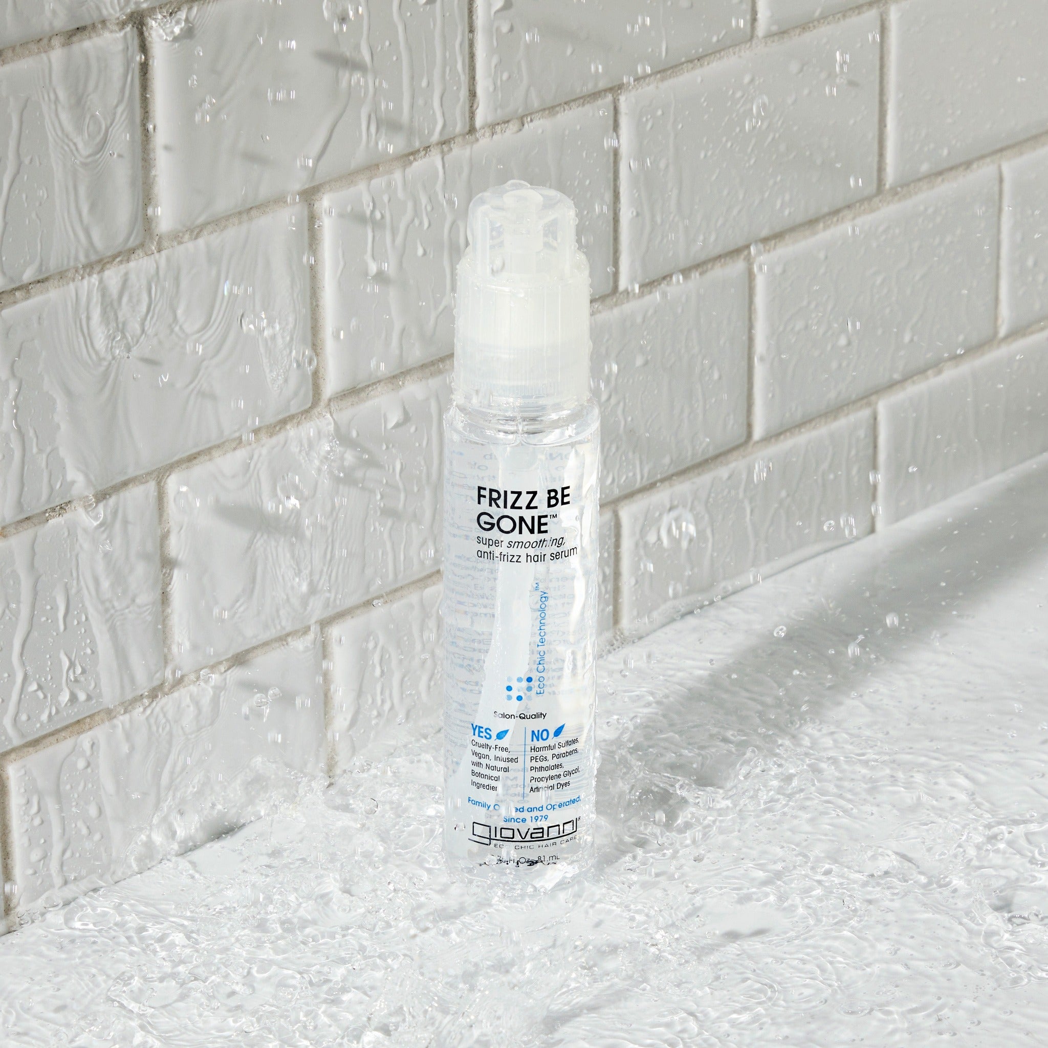 A clear bottle of Giovanni Cosmetics FRIZZ BE GONE™ Super-Smoothing, Anti-Frizz Hair Serum stands upright on a wet, textured surface with water droplets, in front of a white glossy tiled wall with splashes.