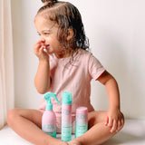 A young child with wet hair, smiling in a pink outfit, sits cross-legged with three turquoise and pink bottles of Giovanni Cosmetics ECO CHIC® KIDS CURL ACTIVATOR SPRAY for moisture retention placed in front of them.